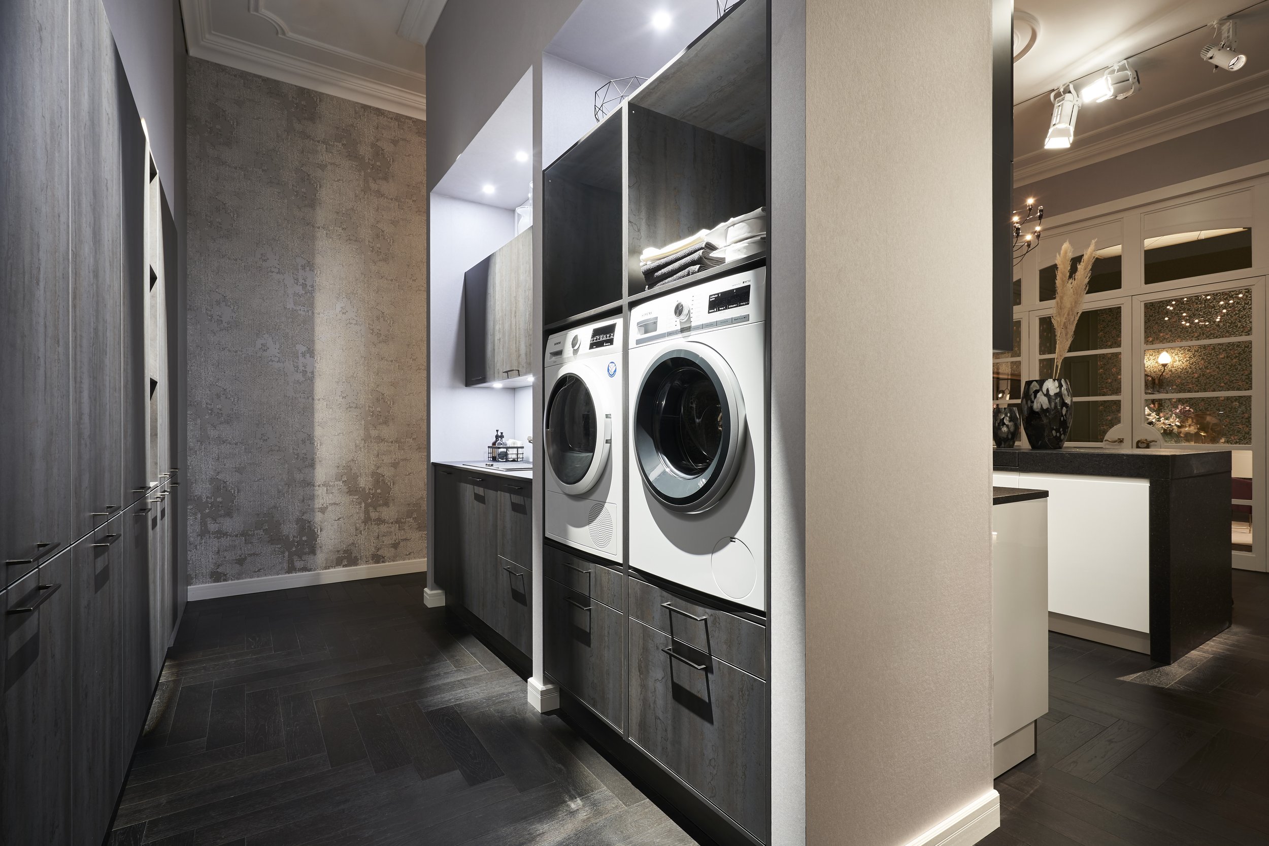 Design laundry space with built-in washer and dryer, dark wood cabinetry, open shelving and soft overhead lighting